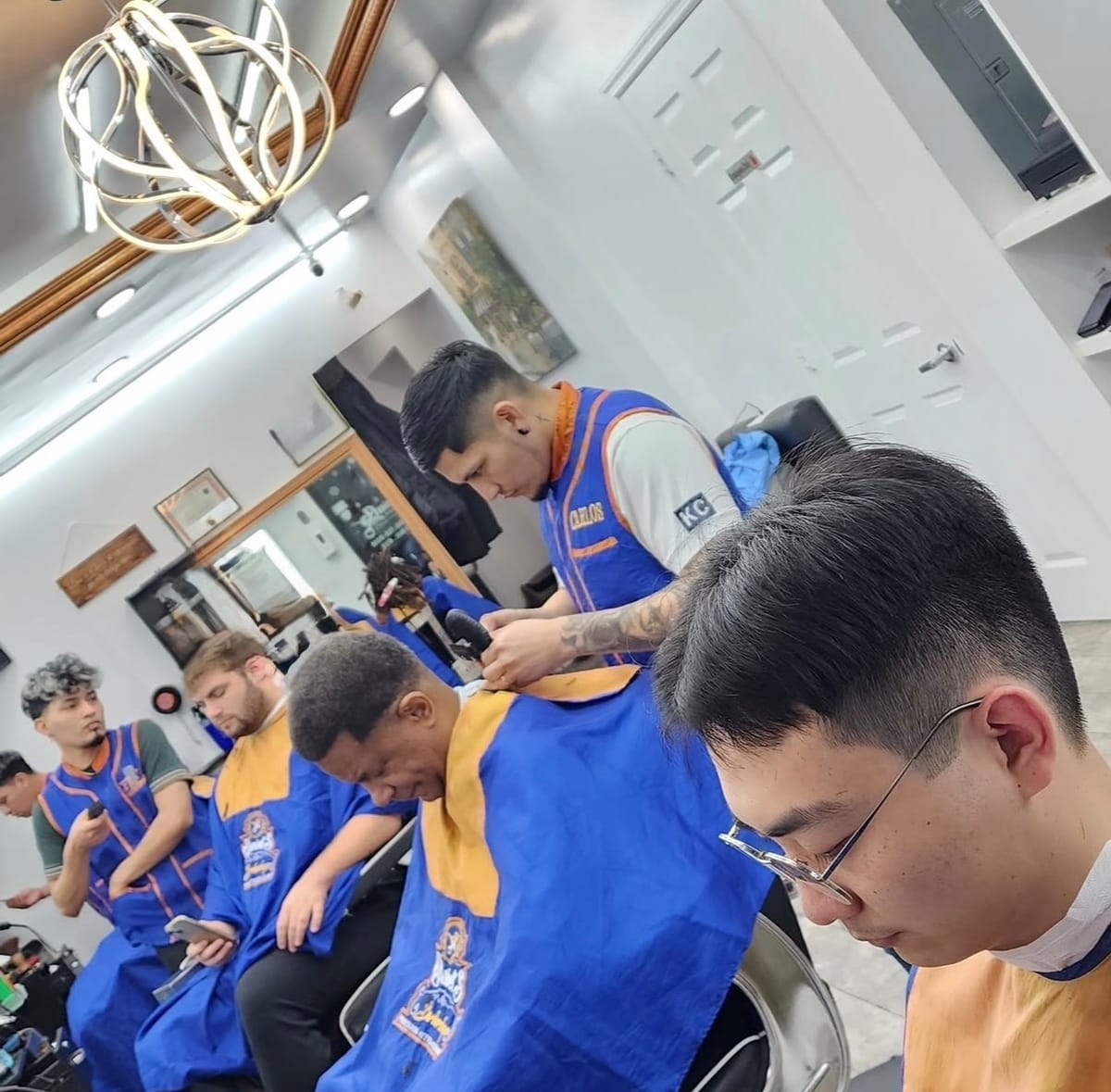 Barbers at work along the row of chairs, clients with their backs to the camera, under the chandelier