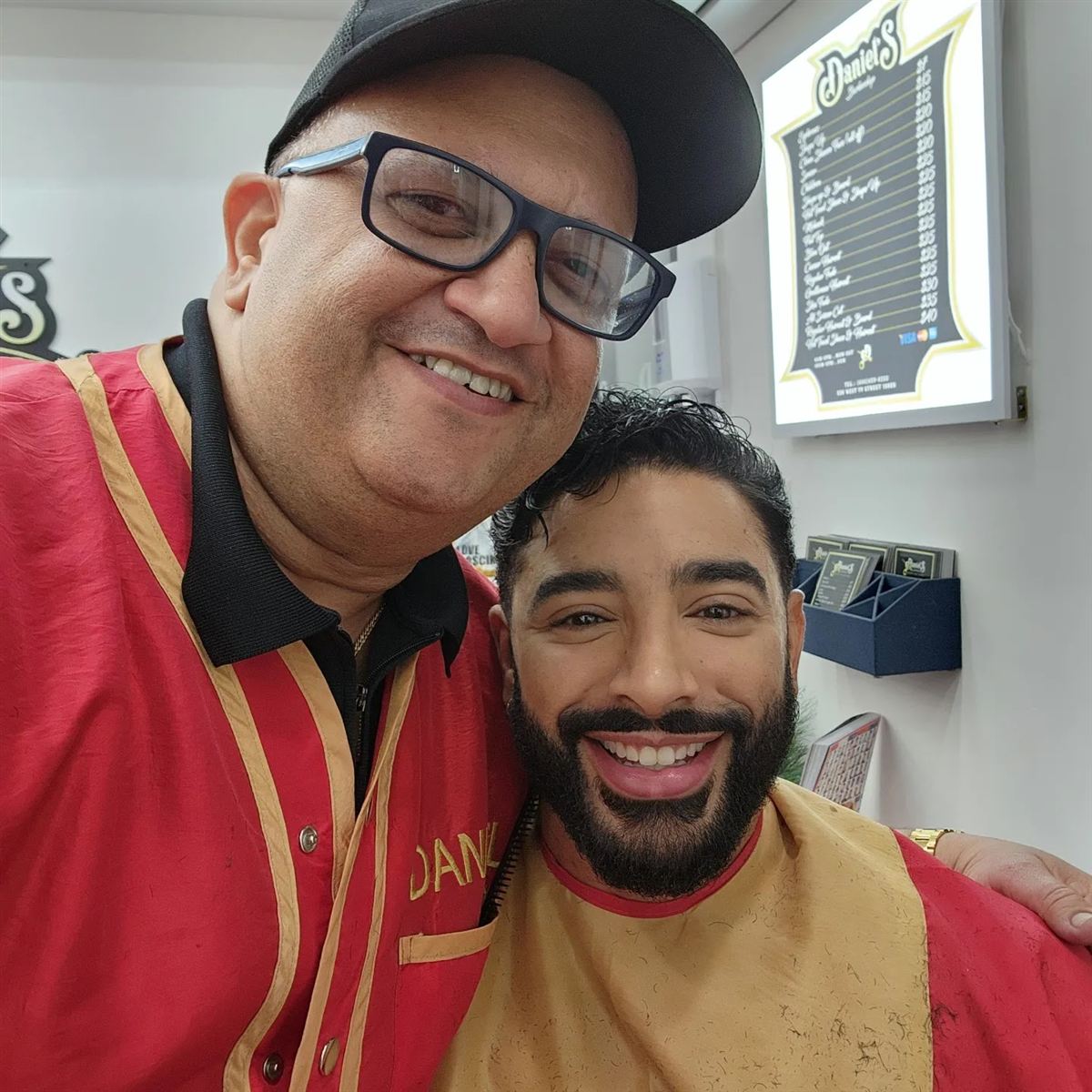 Daniel in his red vest and cap taking a selfie with a smiling client in a gold-collared cape